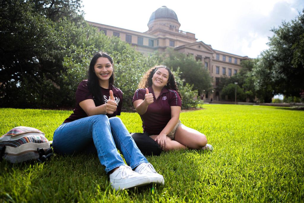 Two Aggie students give thumbs up as they sit on the lawn in front of the Academic Building