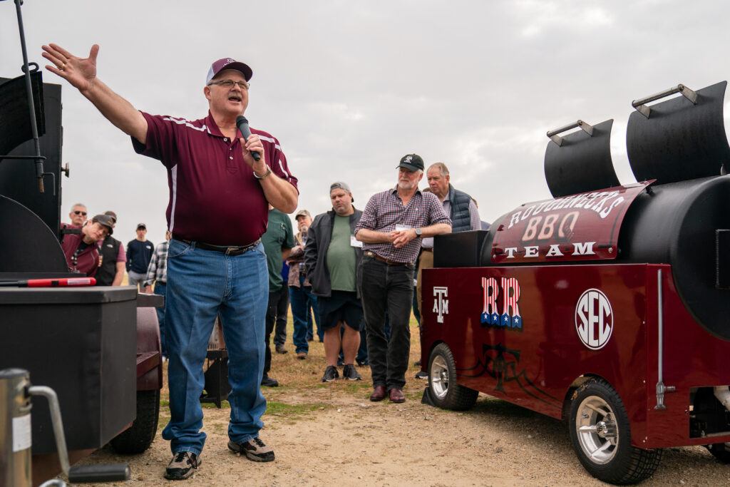 A speaker, Dr. Davey Griffin, stands welcoming barbecue enthusiasts who are gathered outside around barbecue pits.