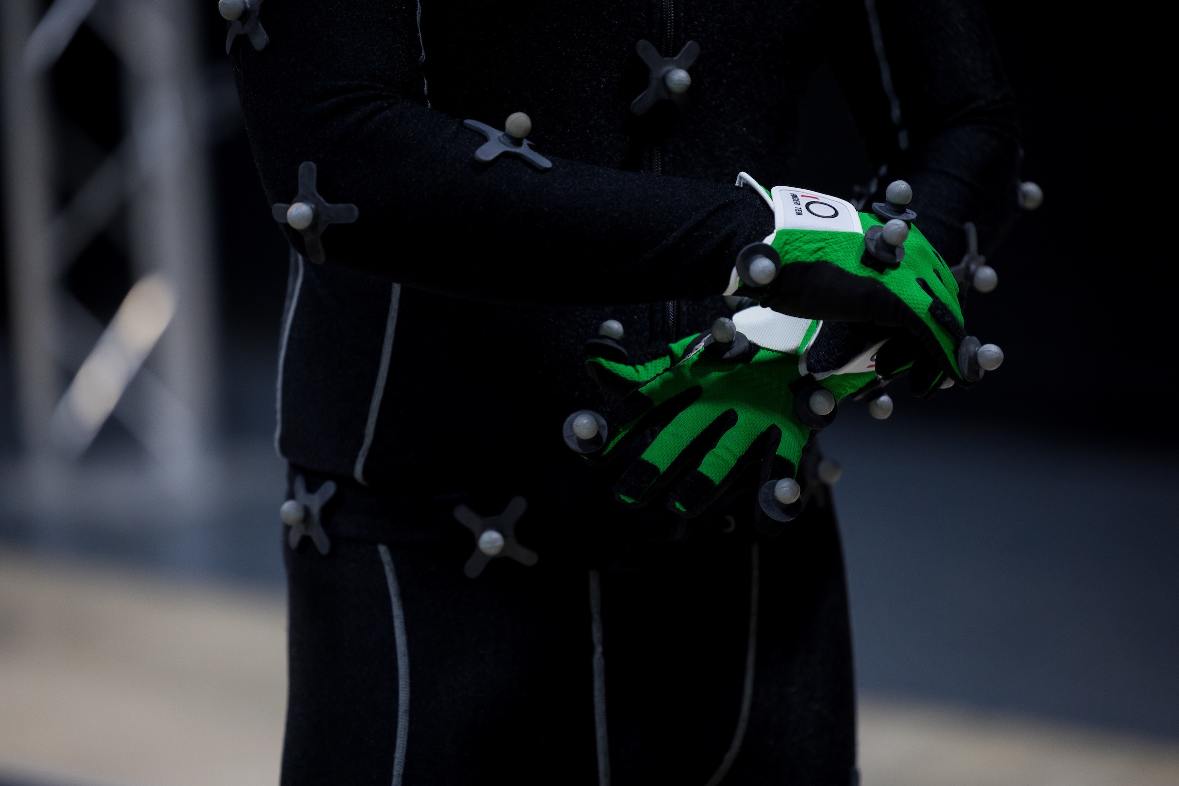 a close-up of a man in a black motion capture suit with green gloves clasping his hands together. The gloves are covered in gray spheres that serve as motion capture markers.