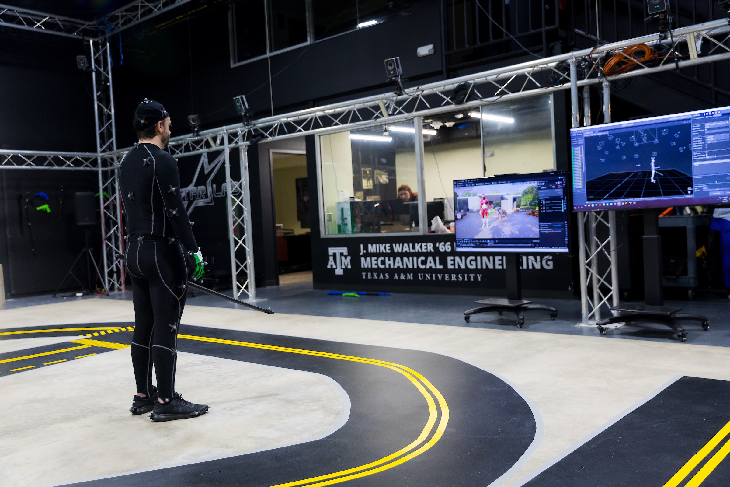 a man in a black motion capture suit covered in grey markers stands in front of two large monitors showing his digital avatar