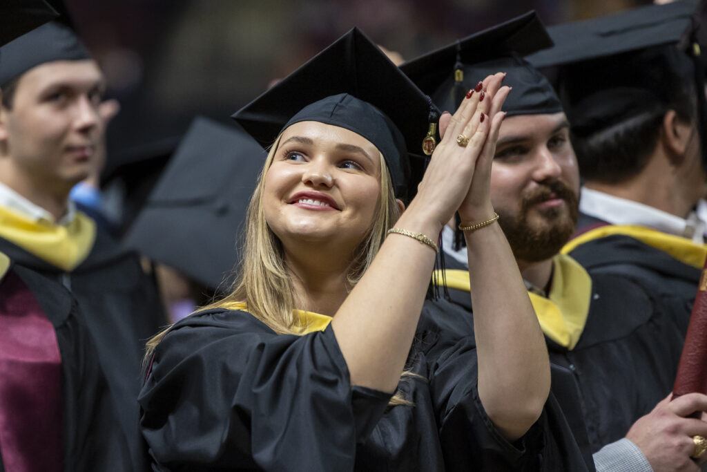 A graduate in cap and gown is surrounded by other graduates and claps while looking toward family and friends in the audience during a commencement ceremony.