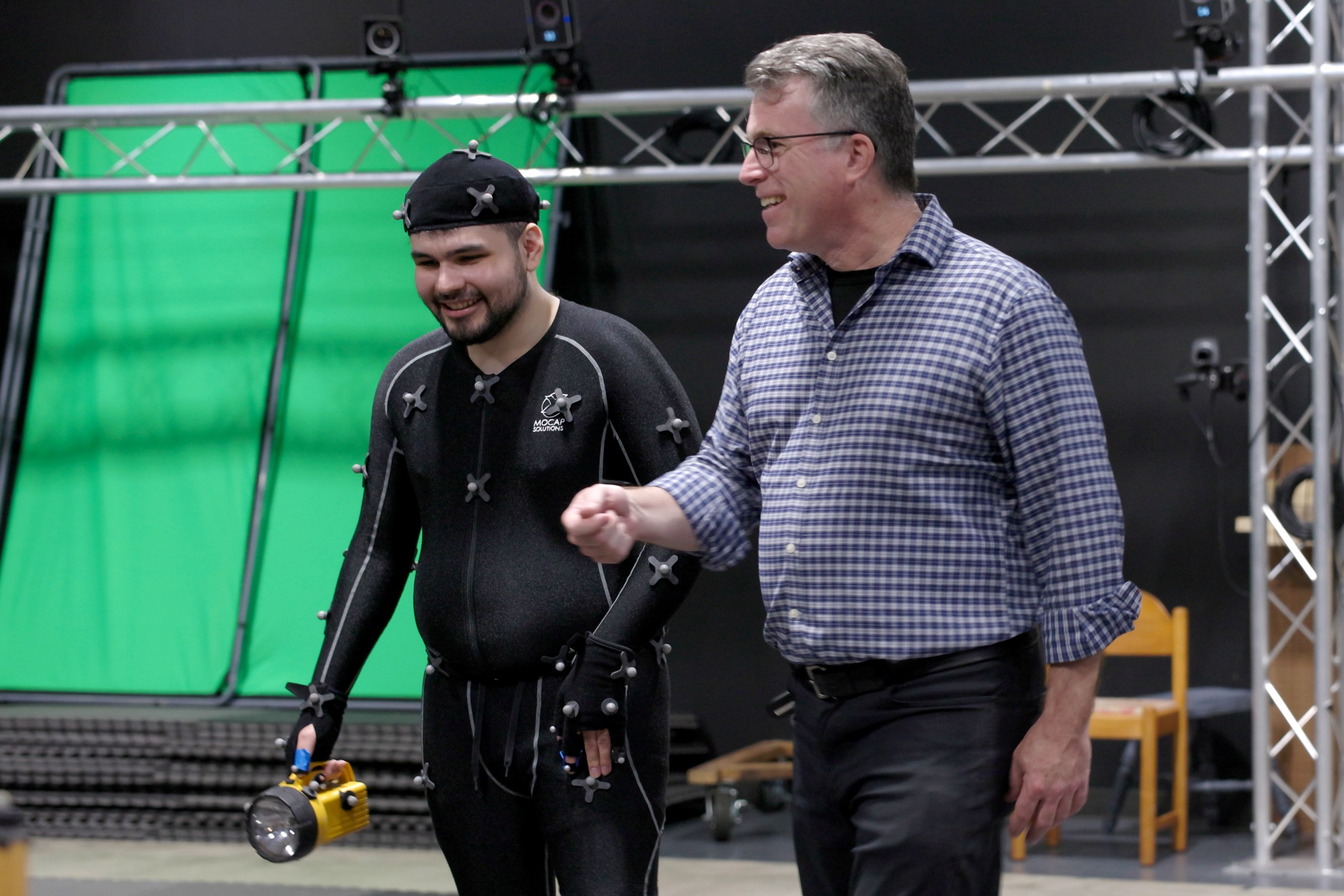 a photo of a man in a checkered shirt and a younger man in a black motion capture suit on a motion capture stage with cameras and a greenscreen in the background