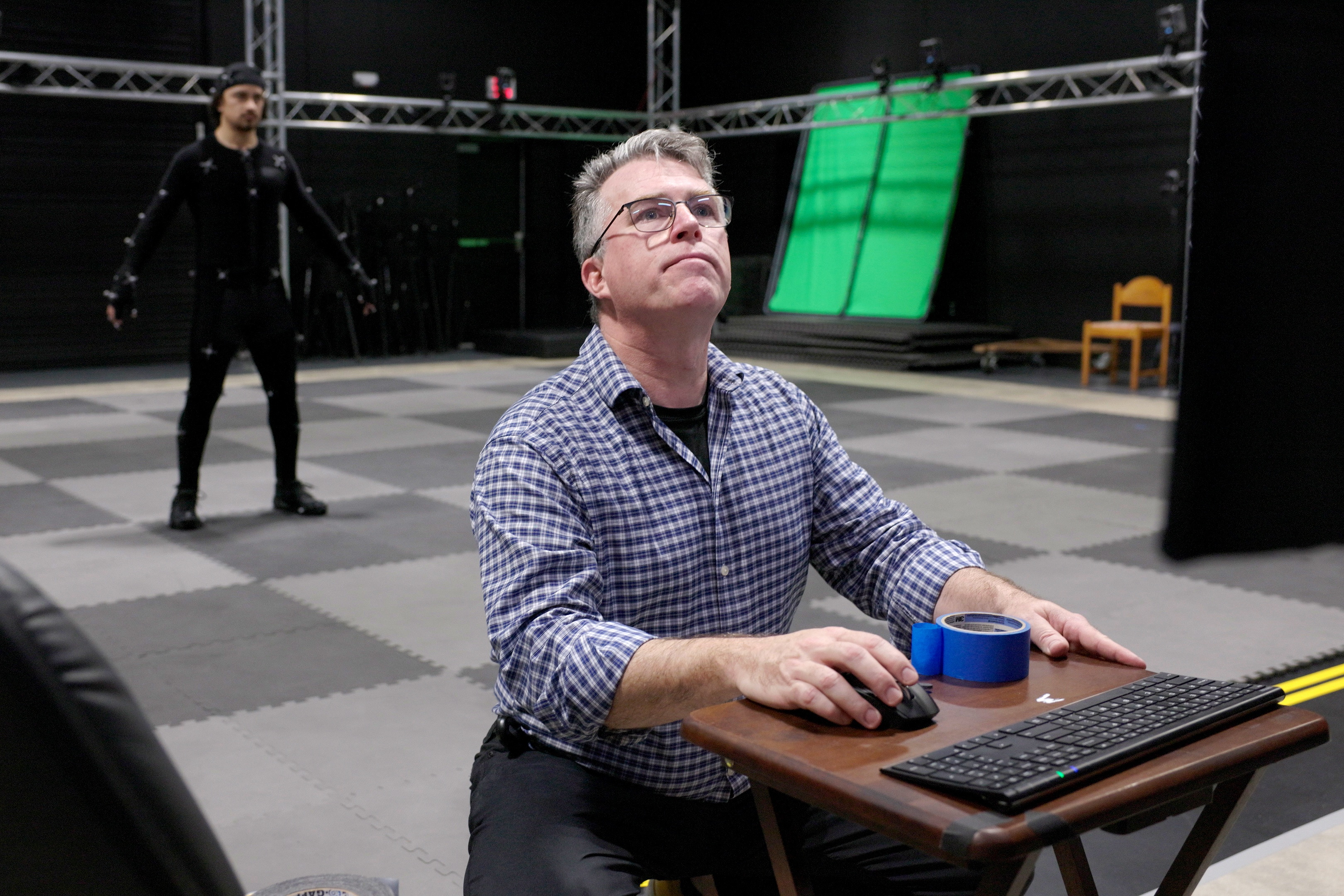 A photo of a man seated at a computer with motion capture stage behind him. A performer in a black motion capture suit stands in a neutral pose near the center of the stage.