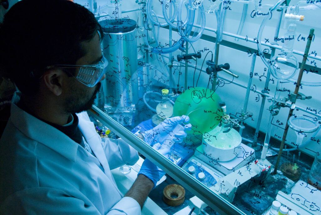 A researcher wearing a lab coat and goggles standing at a lab bench