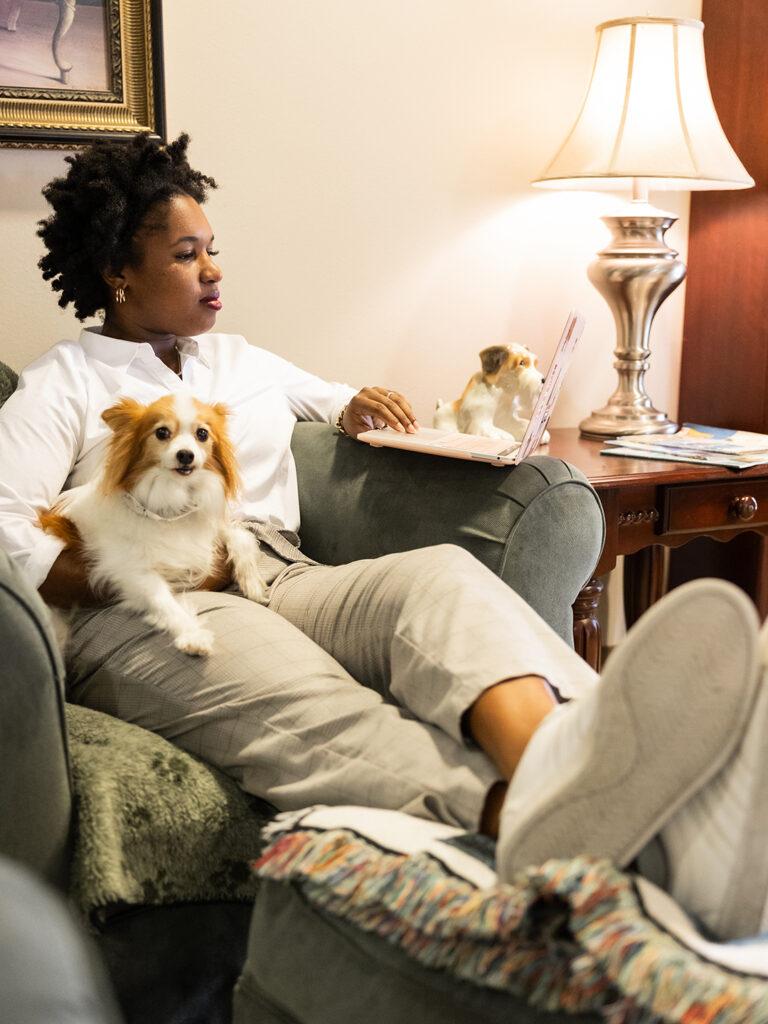 A woman sitting in a chair with a dog on her lap while looking at a laptop computer.