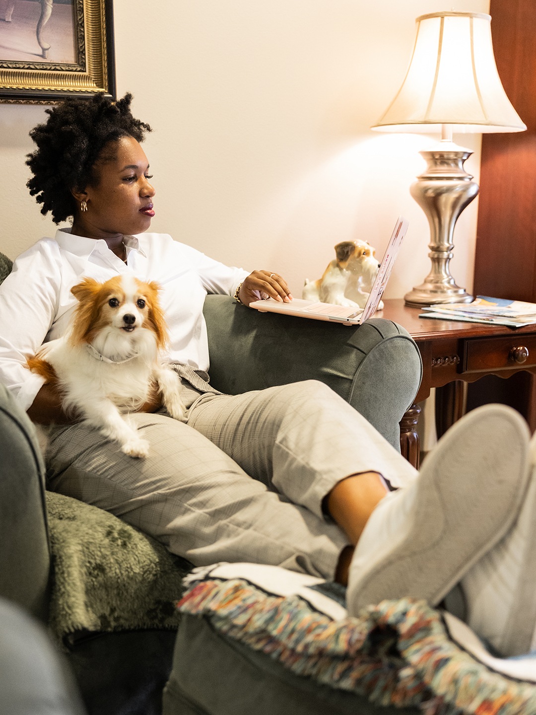 A woman sitting in a chair with a dog on her lap while looking at a laptop computer.