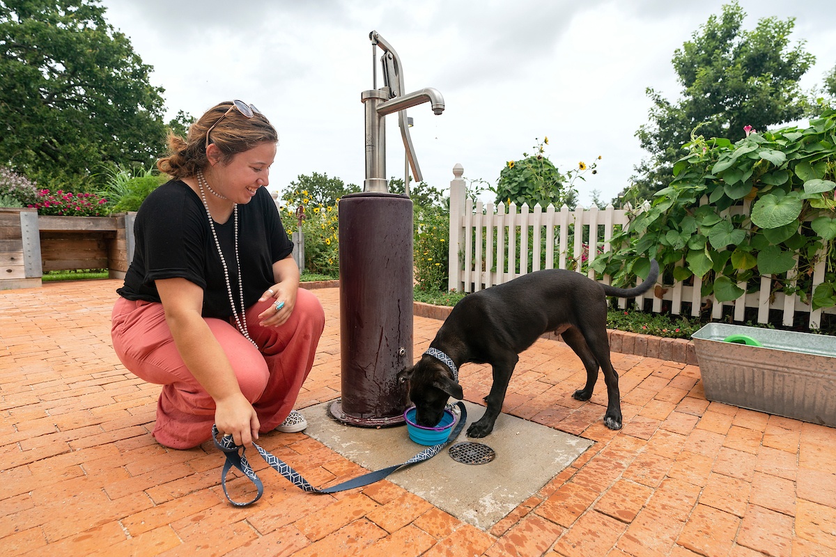 A woman squats next to a water pump. She is holding a dog leash while her dog drinks from a bowl. The dog is black in color and she is wearing a black top and pink pants. There are trees and a white picket fence behind them