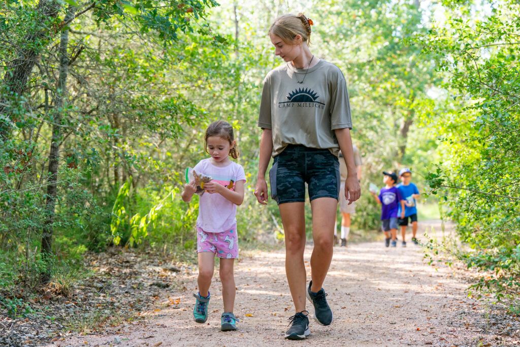 A woman and a young girl walk down a dirt path through a wooded area with some young kids behind them in the distance.