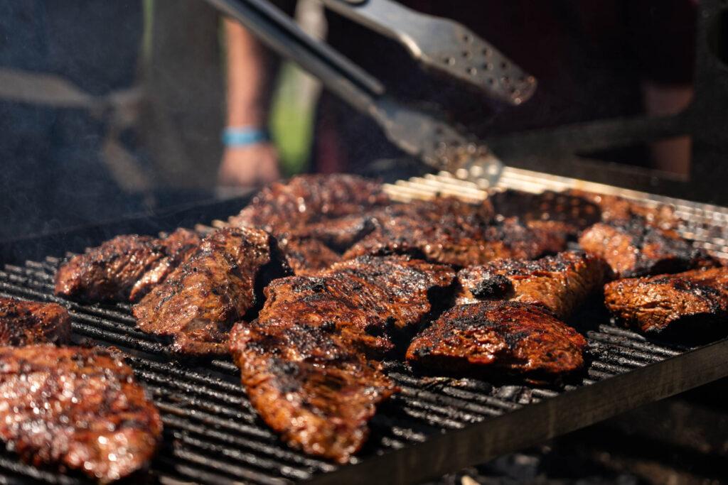 A close-up shot of meat cooking on a grill with someone holding a pair of tongs