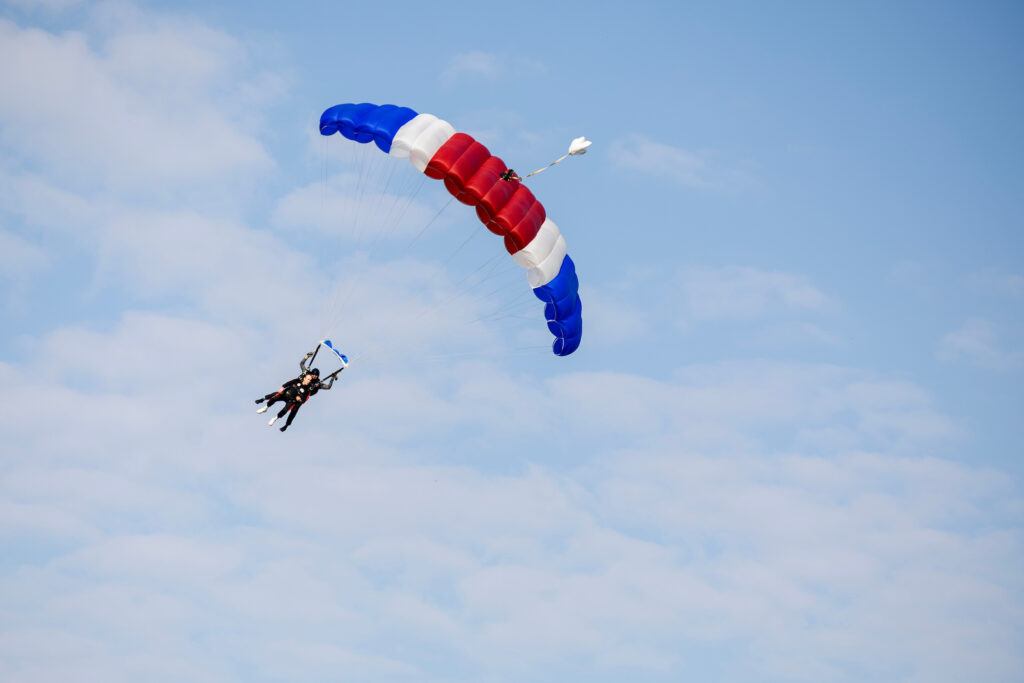 two men hang from a red white and blue parachute with clouds in the background