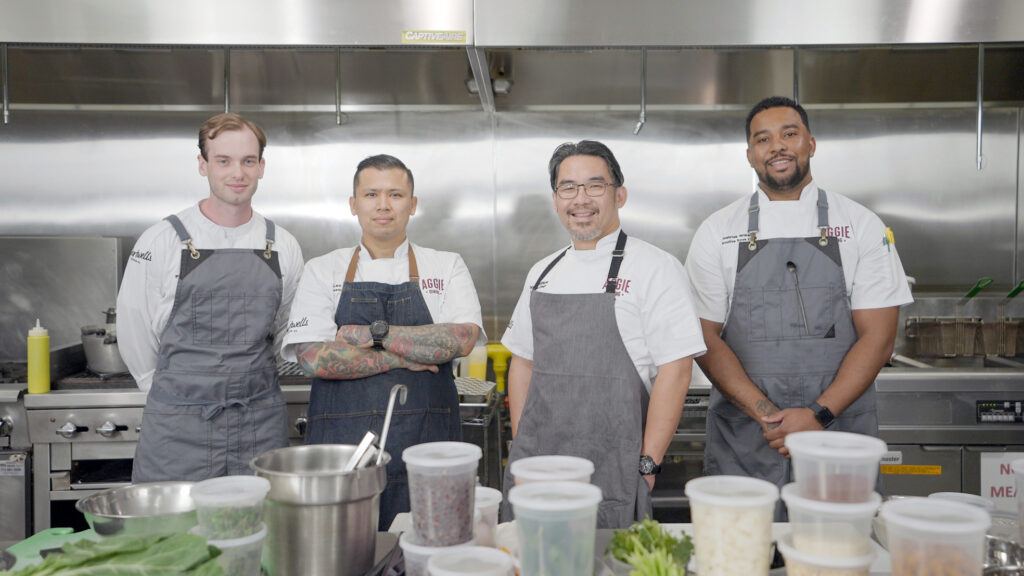 four men in aprons stand behind a counter loaded down with various cooking ingredients