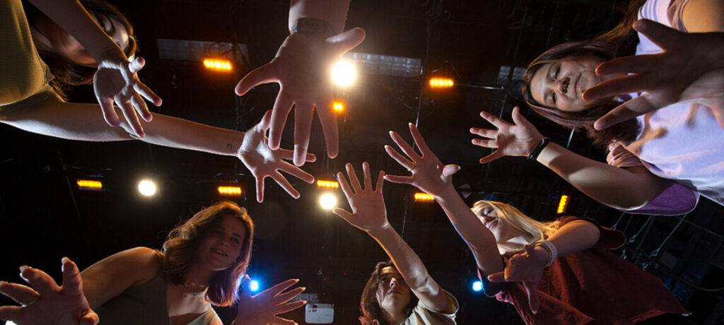 A group of theater students puts their hands together prior to a performance under theater lights.