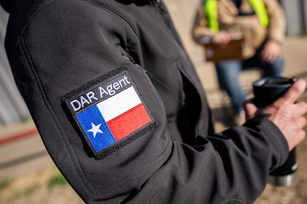 The right arm of an individual wearing a jacket with a Disaster Assessment and Recovery badge on it. The individual is also holding a cup in their right hand.