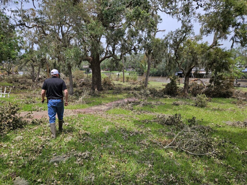 a man wearing jeans and rubber boots walks through a field that has fallen limbs and tree branches.