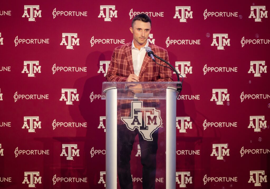 a photo of a man in a plaid suit jacket standing behind a glass podium in front of a maroon Texas A&M backdrop