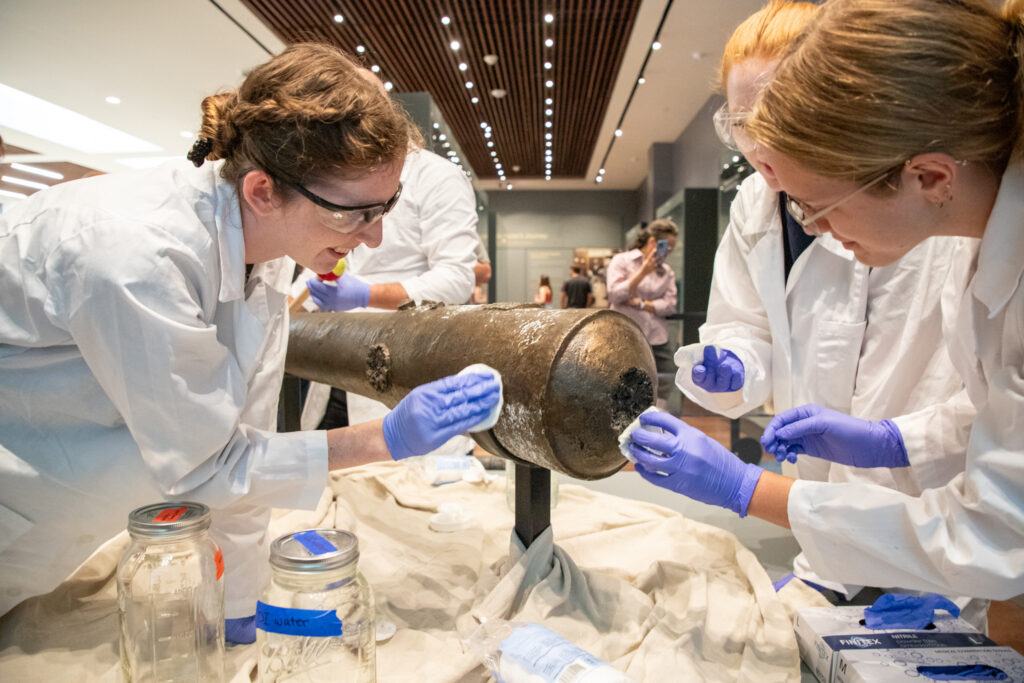 Nautical Archaeology Program graduate students Kimberly Breyfogle '23 (left), Alyssa Carpenter '22 (center) and Marissa Agerton '25 (right) gently clean the exterior of a four-pound bronze cannon used in the Battle of the Alamo.