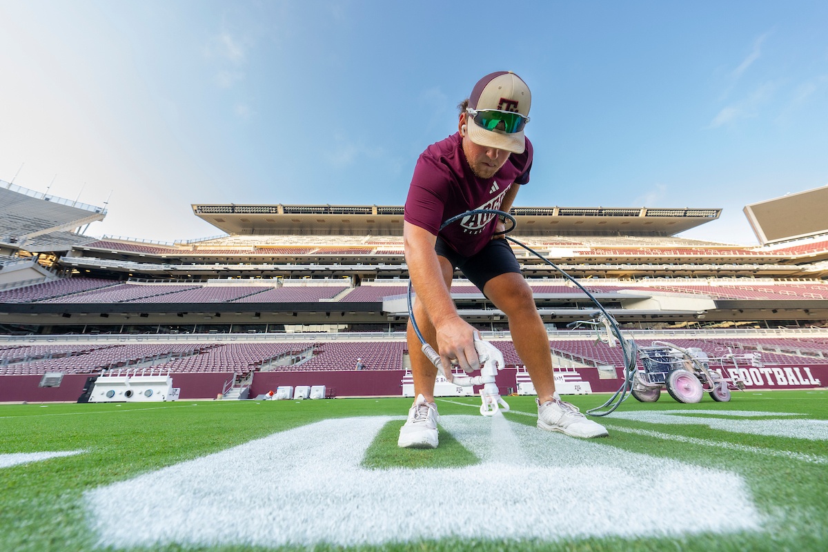 A person in a maroon shirt and cap paints lines on a football field at an empty stadium. He is crouched down, holding a paint sprayer, ensuring the markings are clear and precise.