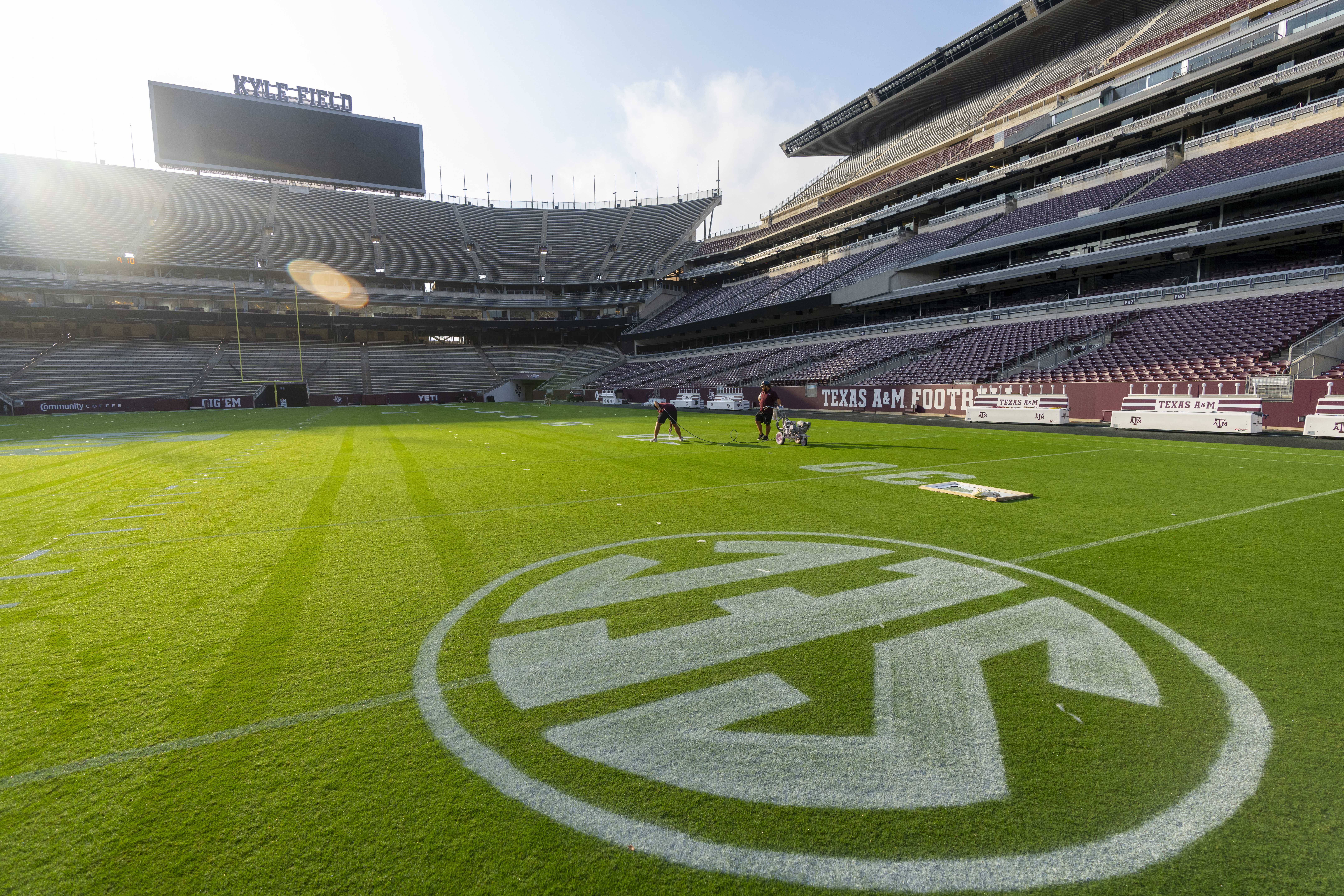 A wide shot of the grass playing surface at Kyle Field with a painted SEC logo in the foreground and two students operating painting equipment in the background