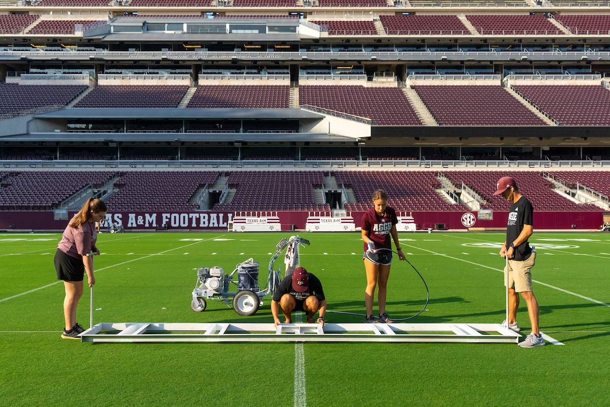 Four turfgrass science students set up equipment on a football field, preparing for line painting. The stadium seats are empty, and "Texas A&M Football" is visible on the stands.