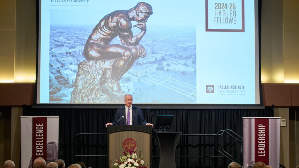 A photo of a man at a lectern on a stage with a video screen behind him during a presentation to an audience.