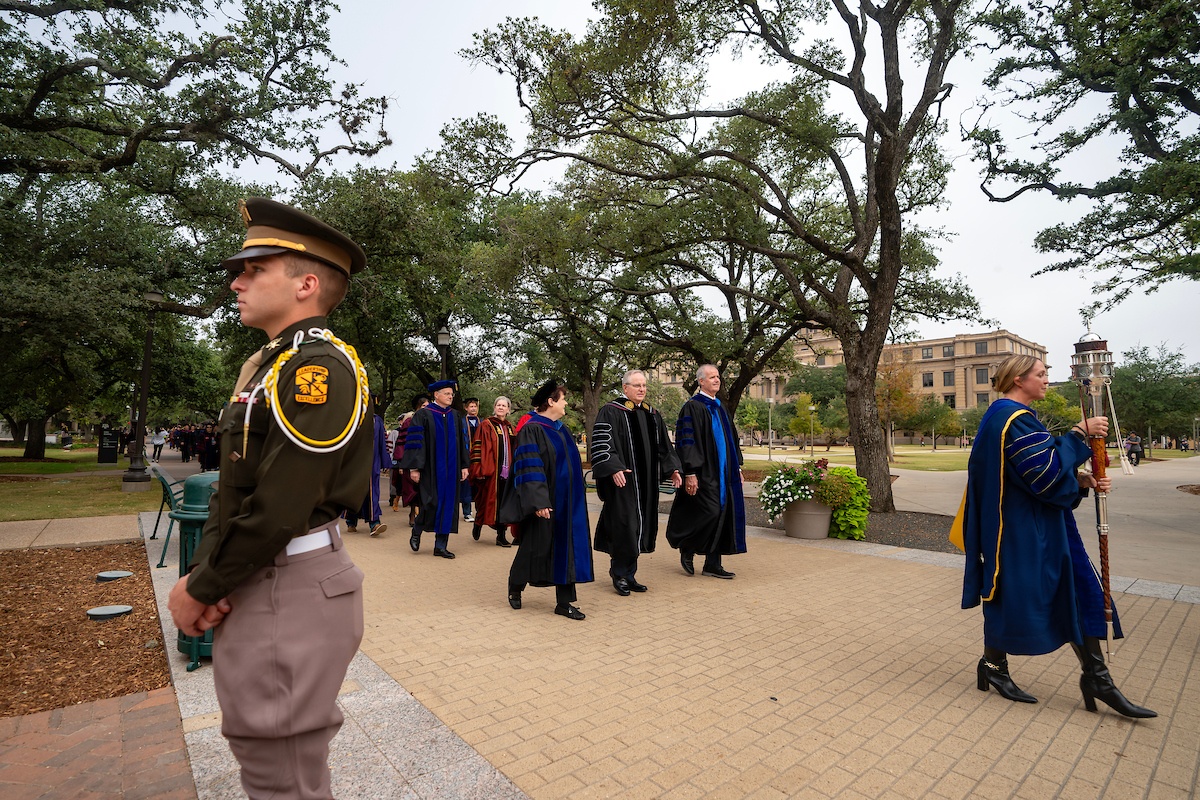 A cadet stands guard as Welsh and other faculty members in regalia walk through campus to Rudder Auditorium