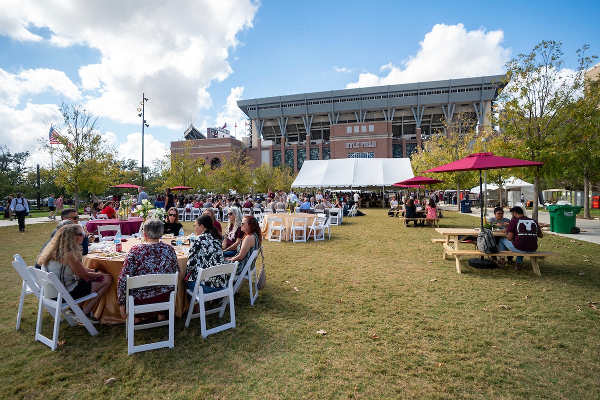 People eat at tables set up outdoors at Aggie Park with Kyle Field in the background