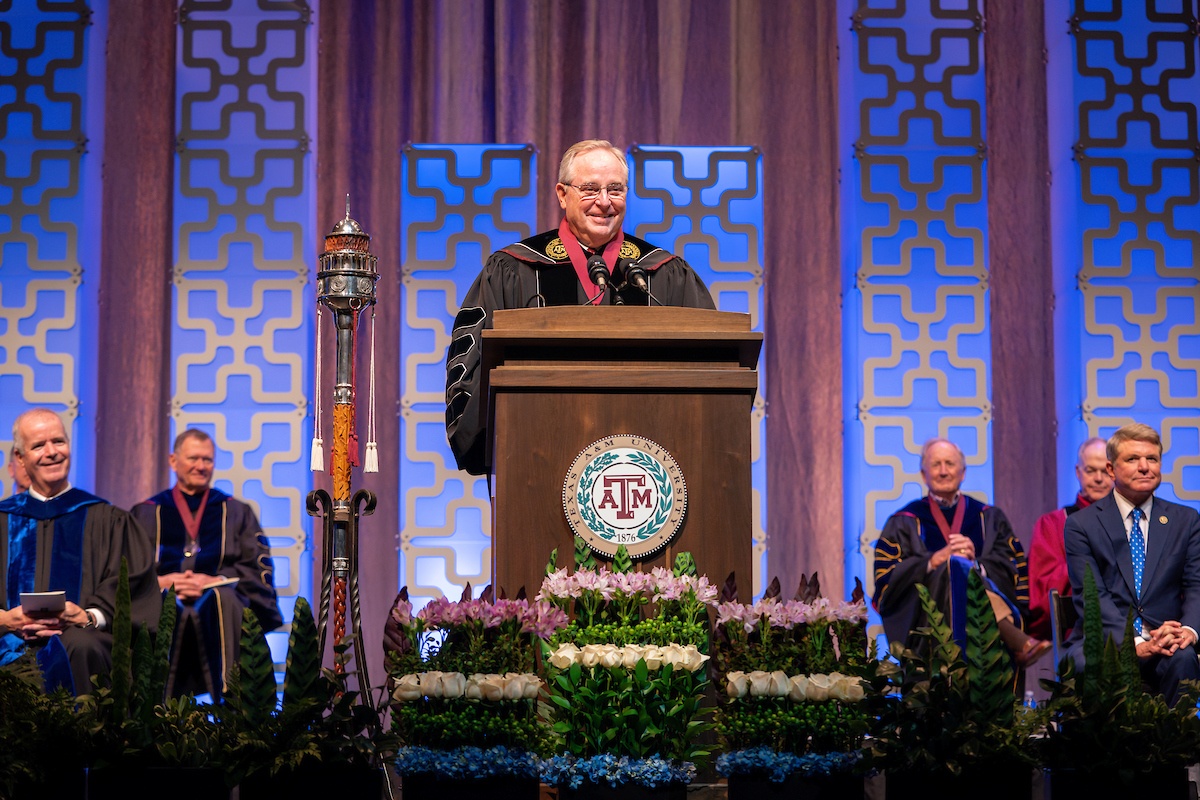 Mark Welsh standing on stage behind a podium as other faculty members look on