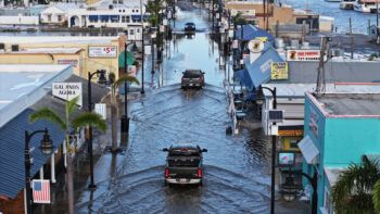 Vehicles drive down a flooded road lined on either side by businesses