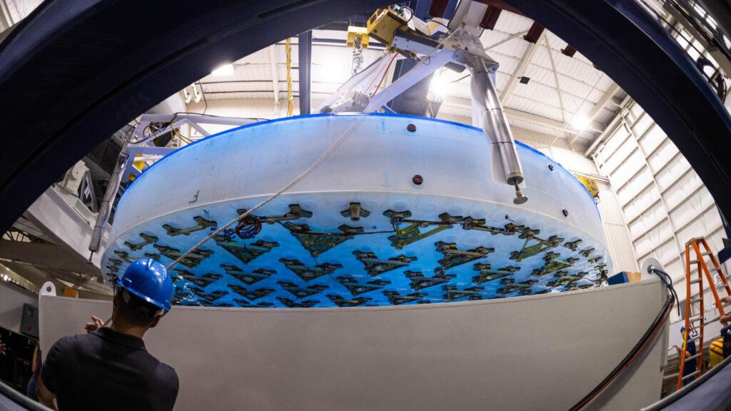 Completed 8.4-meter-diameter primary mirror, hanging above the support system prototype with a technician in the foreground at the University of Arizona’s Richard F. Caris Mirror Lab.