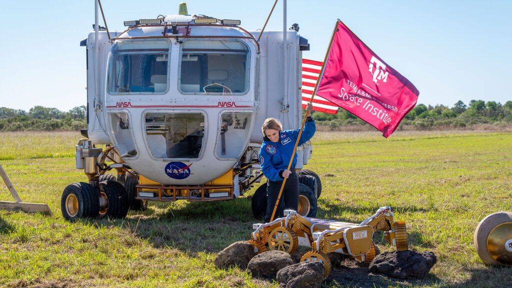 A photo of a woman planting a Texas A&M University flag in a field of grass with NASA vehicles around her.