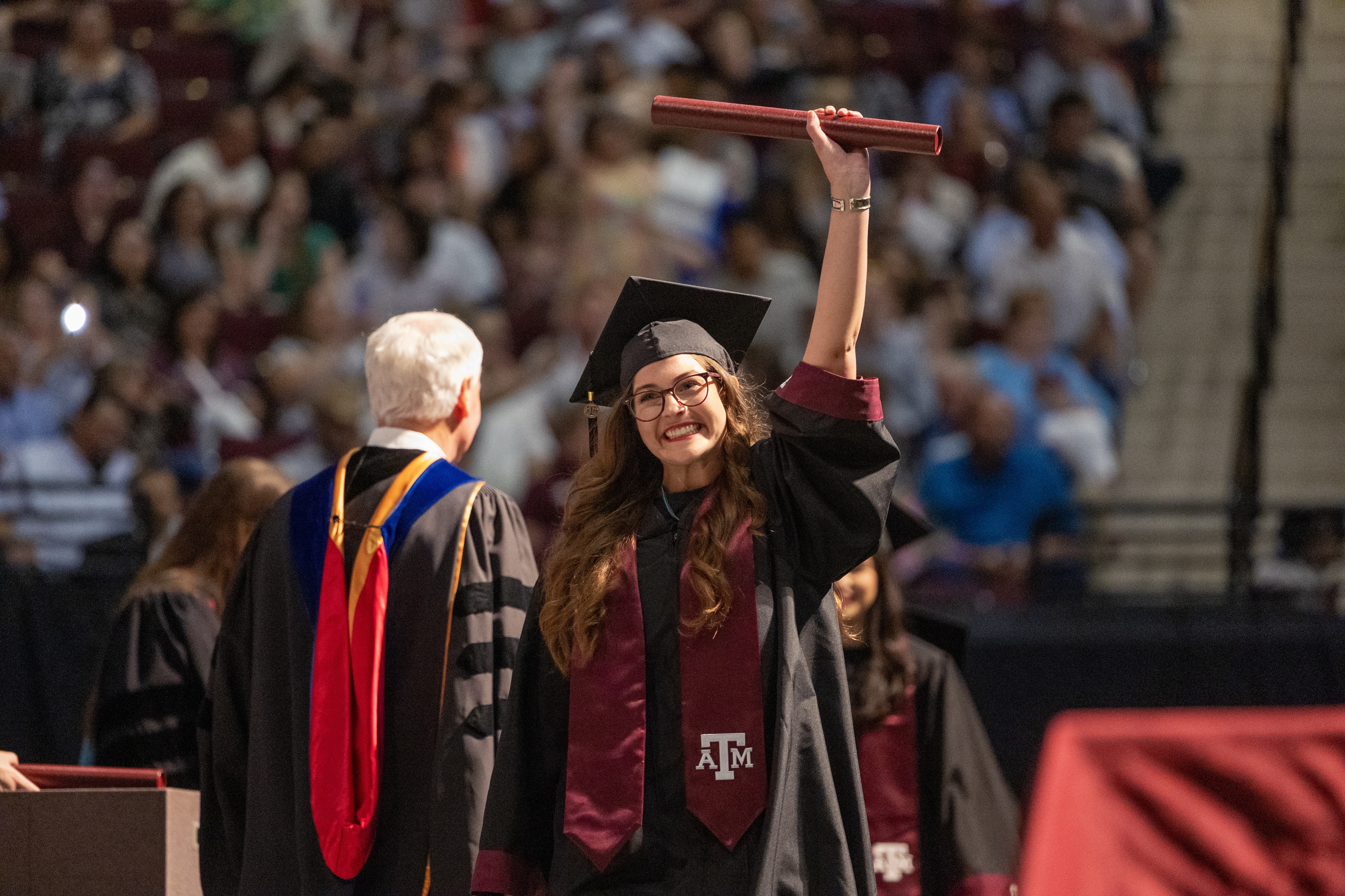 A photo of a graduate holding a diploma tube in the air and smiling to the crowd after crossing the stage during a commencement ceremony.