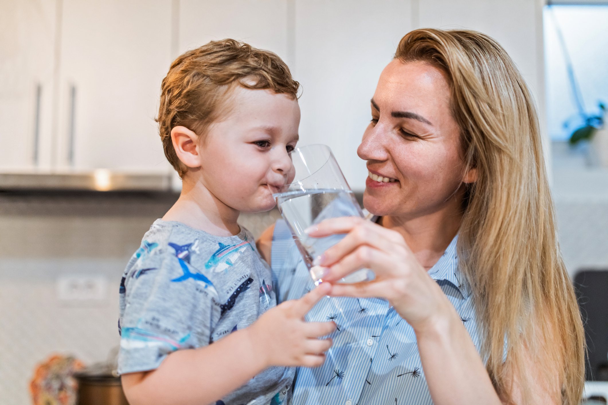 Mother offering a glass of water to her toddler son. Holding a big glass to his mouth.