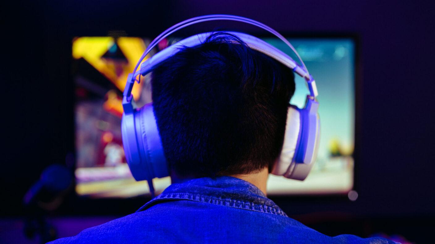A man from behind. He is sitting at a desk wearing a large pair of headphones while a monitor glows on the desk in front of him. A microphone sits next to the monitor.