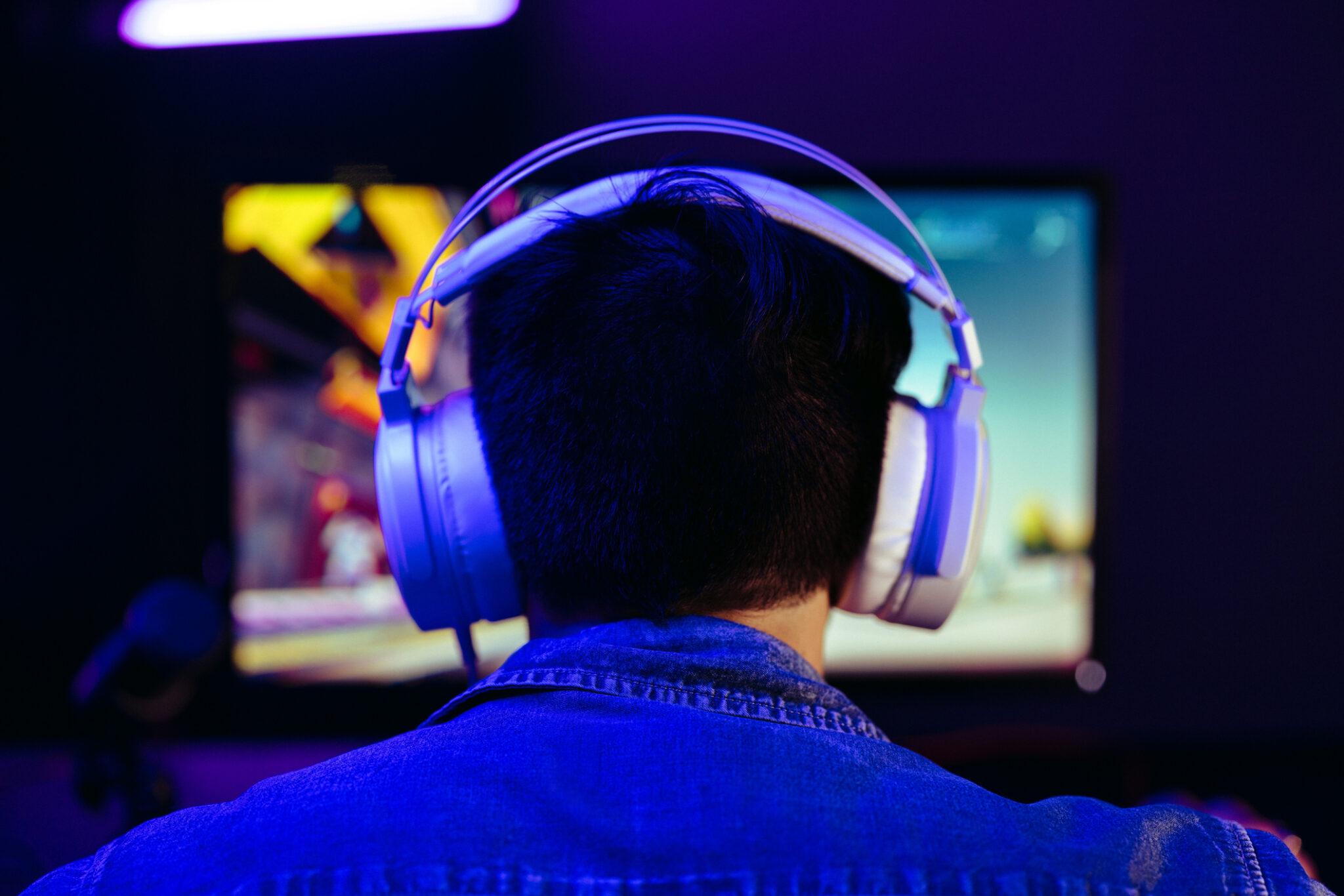 A man from behind. He is sitting at a desk wearing a large pair of headphones while a monitor glows on the desk in front of him. A microphone sits next to the monitor.