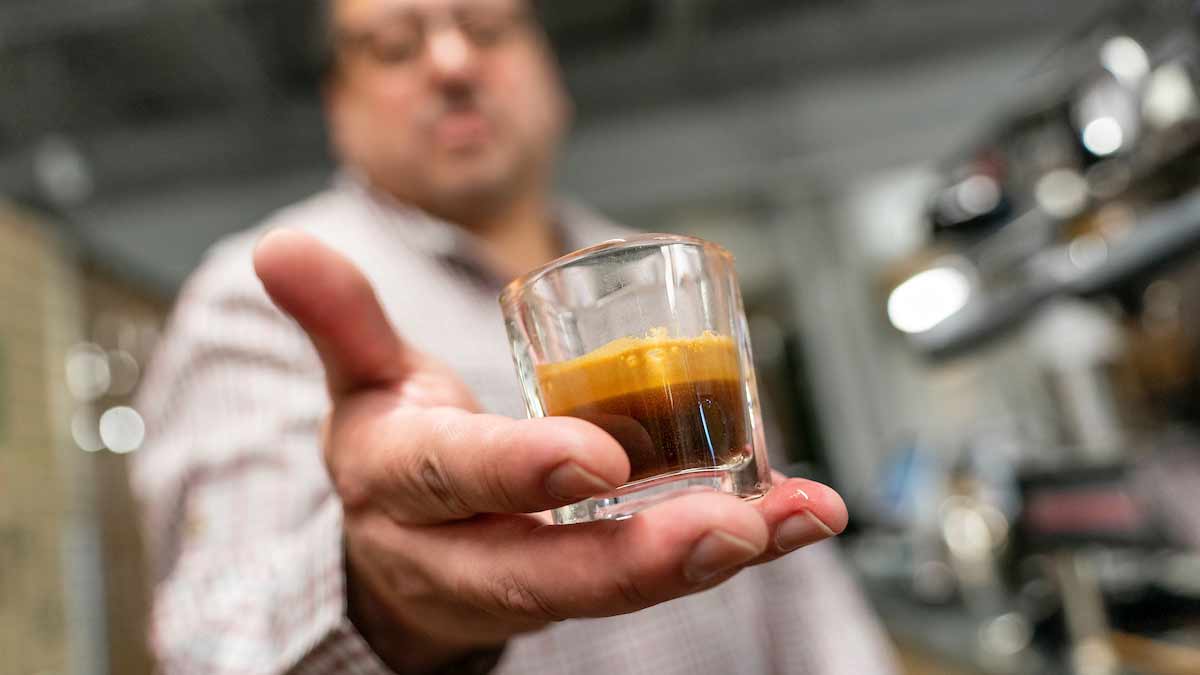 A man in a white lab coat extends a hand holding a glass shot glass filled with espresso toward the camera