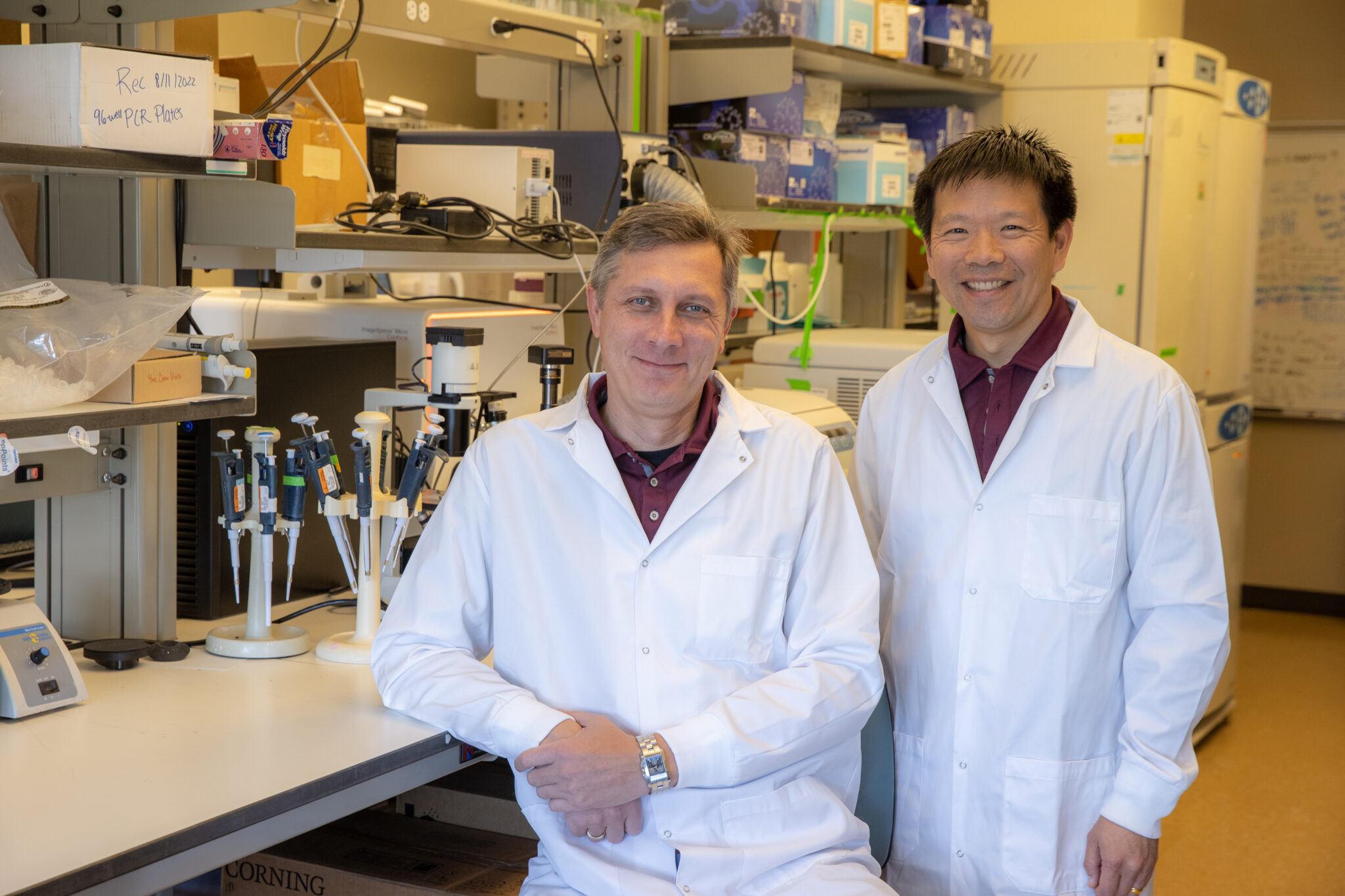 two researchers posing for a photo in a lab while wearing white lab coats.