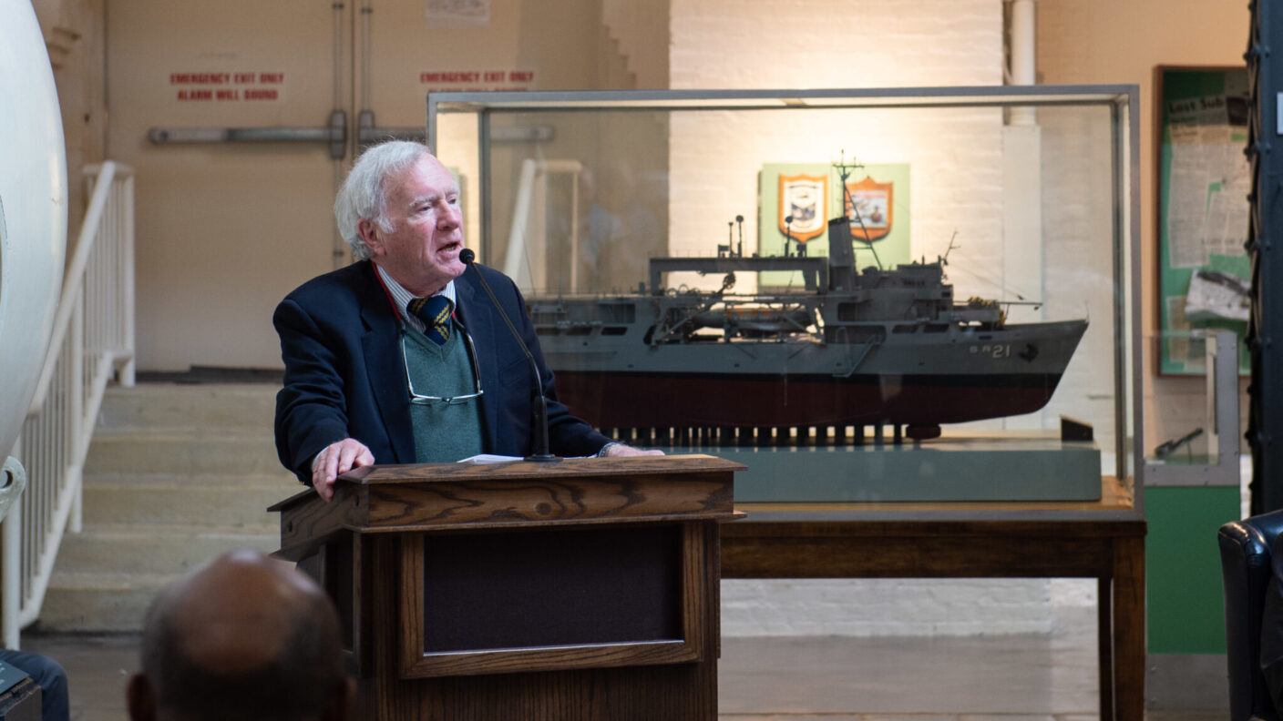 A photo of a man speaking at a lectern with a model of a ship behind him.