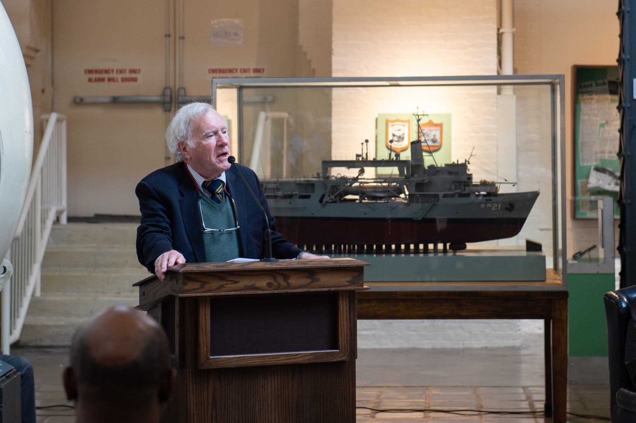 A photo of a man speaking at a lectern with a model of a ship behind him.