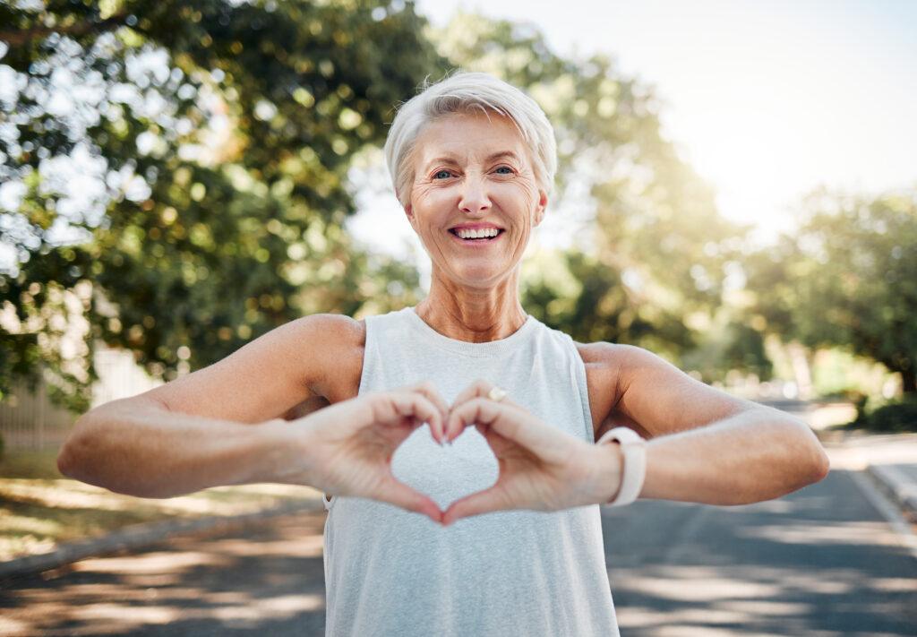 A woman holds her hands together in the shape of a heart while on a walking trail surrounded by trees on a sunny day.