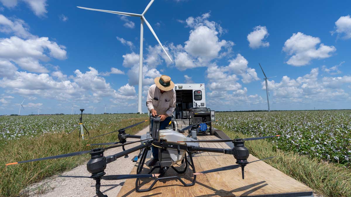 Man in straw hat repairs large drone in the middle of a crop field with wind turbines in the background