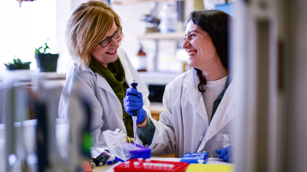 Dr. Shippen wearing a white lab coat smiles and talks to a lab assistant