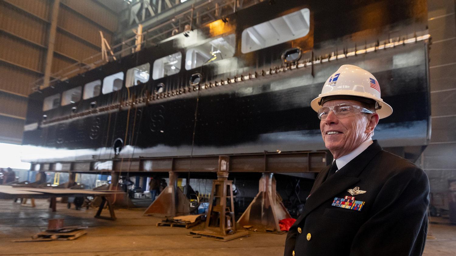 Man in military dress and a hard hat smile in front of ship being built