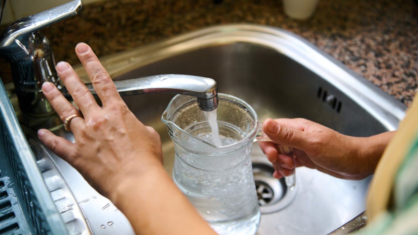 A close-up image of hands filling a glass pitcher with water from a kitchen faucet.