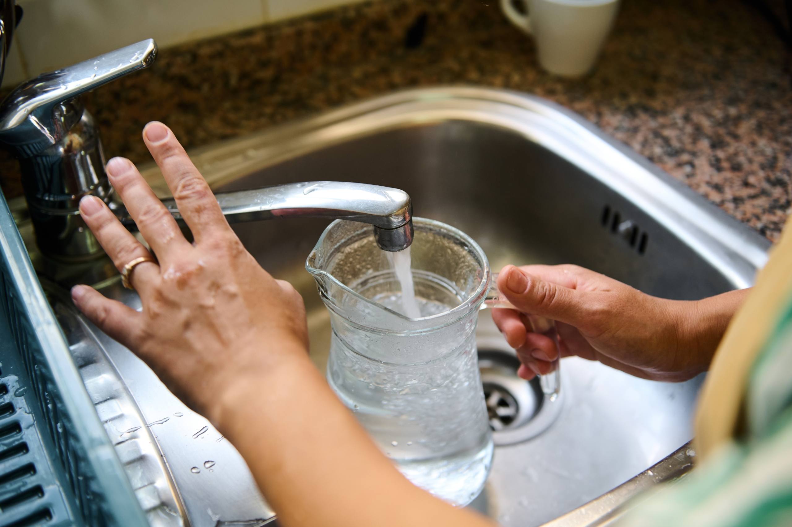 A close-up image of hands filling a glass pitcher with water from a kitchen faucet.