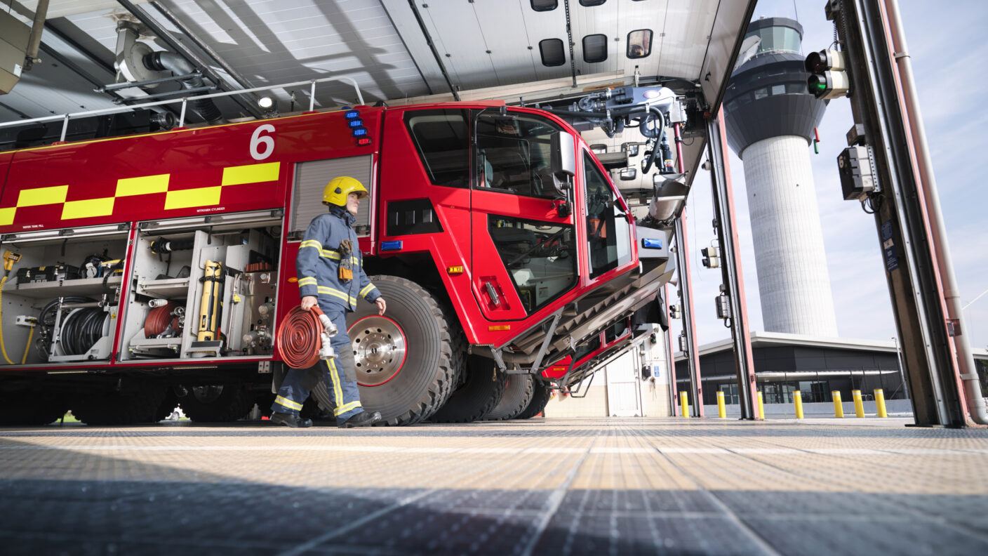 A photo of a firefighter carrying equipment to a fire engine in an airport fire station.