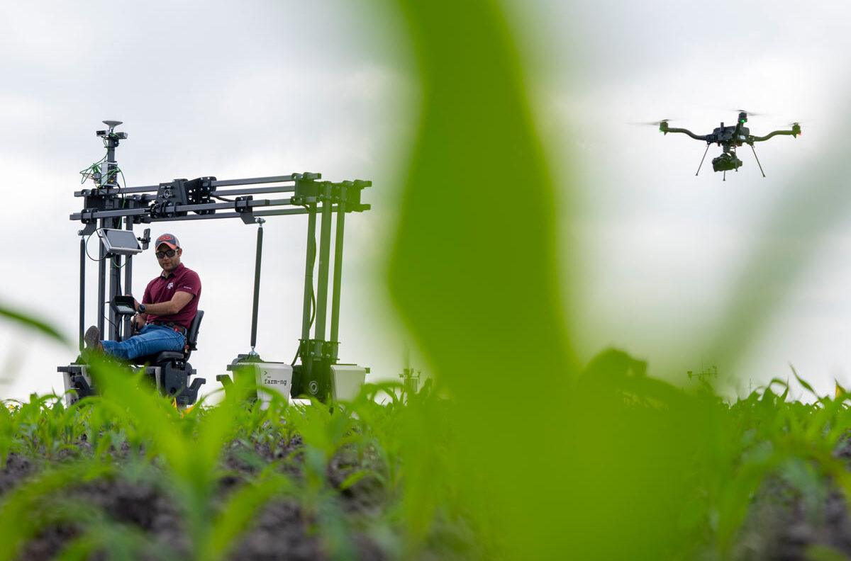 A farmer sits on a a piece of farming technology in a field while a drone flies by