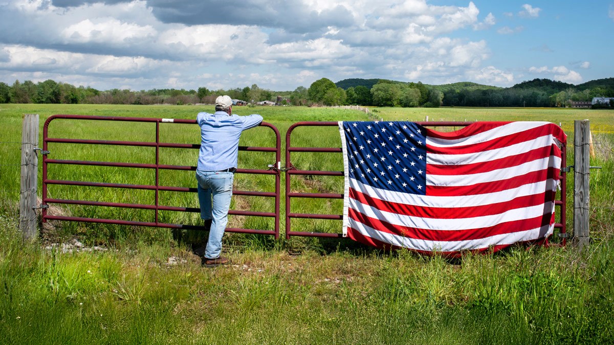 A farmer leans against a gate that is holding an American flag