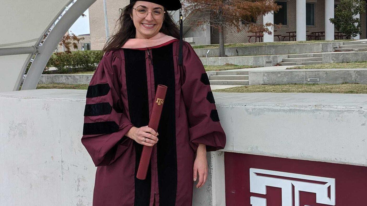 Ashley Hotard standing next to the Texas A&M logo in her graduation cap and gown