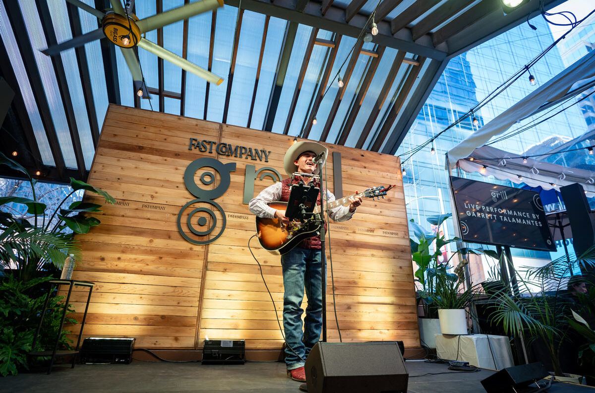 Garrett Talamantes in an Aggie vest and cowboy hat plays acoustic guitar on the Fast Company Grill stage