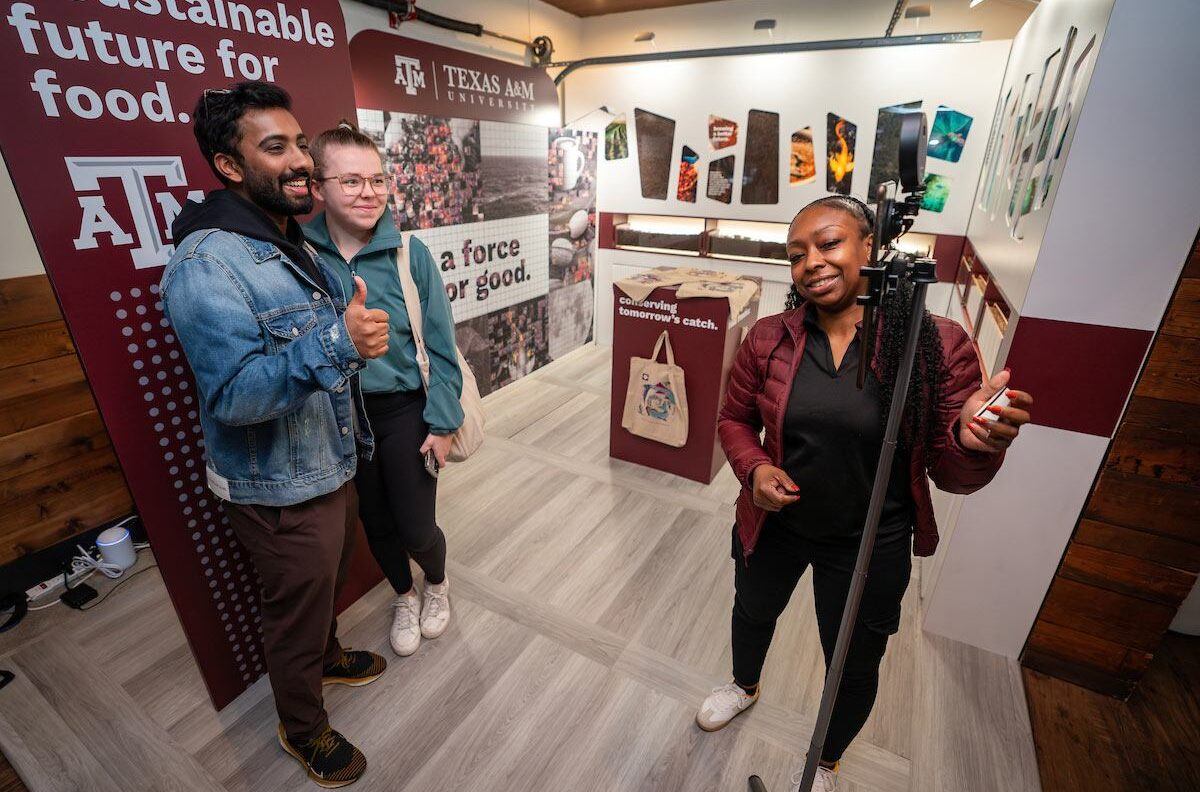 A Texas A&M representative helps people take selfies in front of a Texas A&M banner